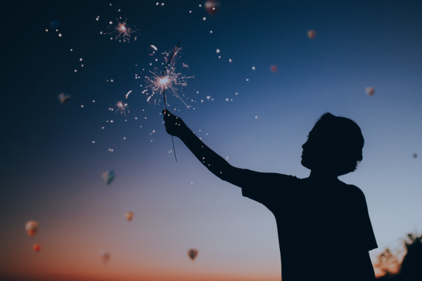 Silhouette of a person holding a lit sparkler against a twilight sky. 