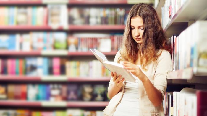 Young woman reading a book leaning against a library shelf.